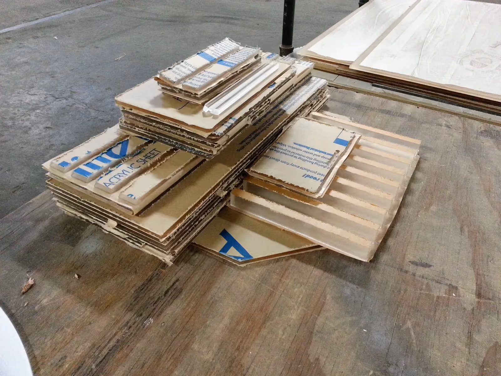 Cut acrylic strips and panels wrapped in brown paper printed Acrylic Sheet, piled on a worn plywood workbench
