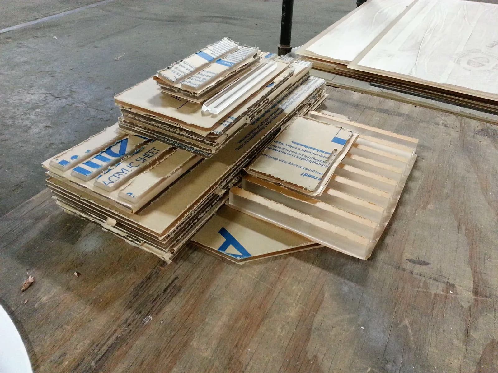 Cut acrylic strips and panels wrapped in brown paper printed Acrylic Sheet, piled on a worn plywood workbench