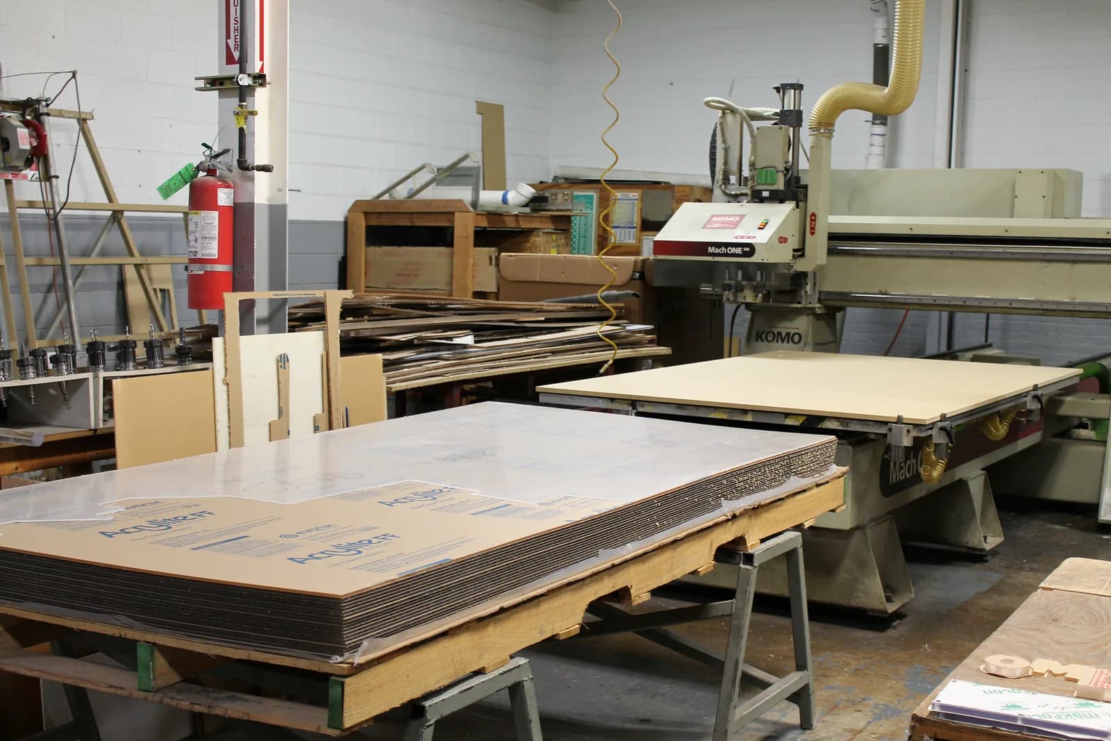 A tall stack of acrylic sheets in protective paper on sawhorses, with a large CNC router and stacked offcuts behind it