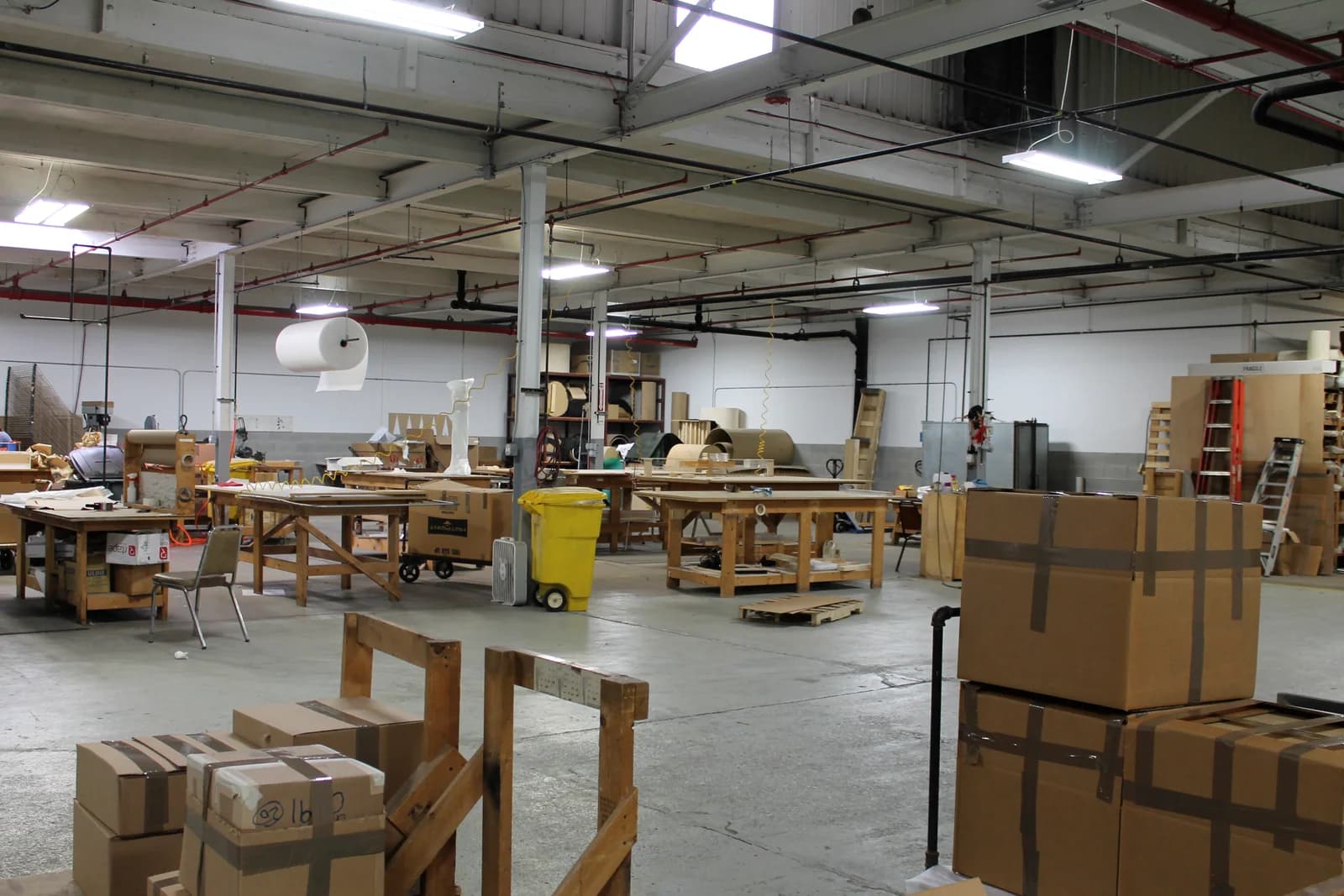 A wide fabrication shop with wooden workbenches, a hanging roll of packing material and shelves of materials, with taped cardboard boxes in front