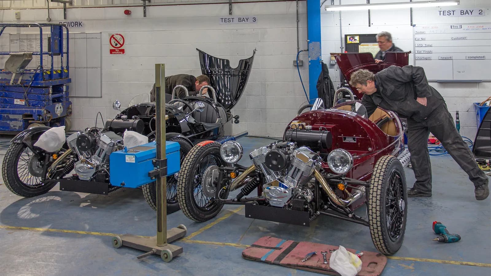 Mechanics work on two open three wheeled roadsters in a factory test bay; boxes mark 3 people.