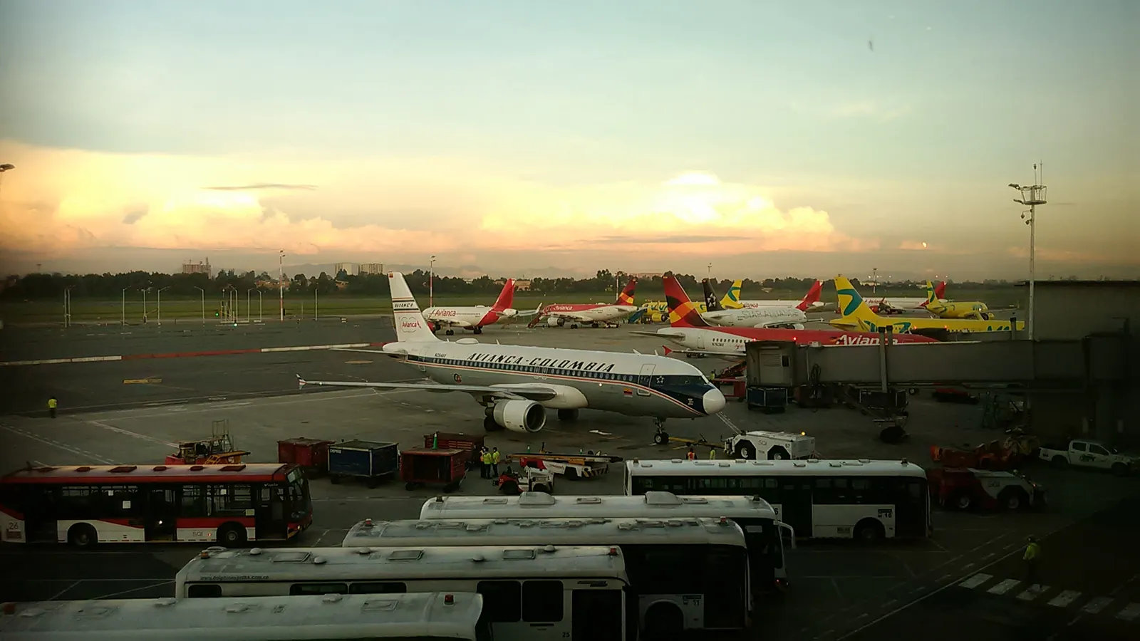 Airliners wait at the gates of El Dorado International Airport in Bogotá at dusk while apron buses line up in front; boxes mark 7 aircraft, 5 buses, 4 service vehicles and 5 ground crew.