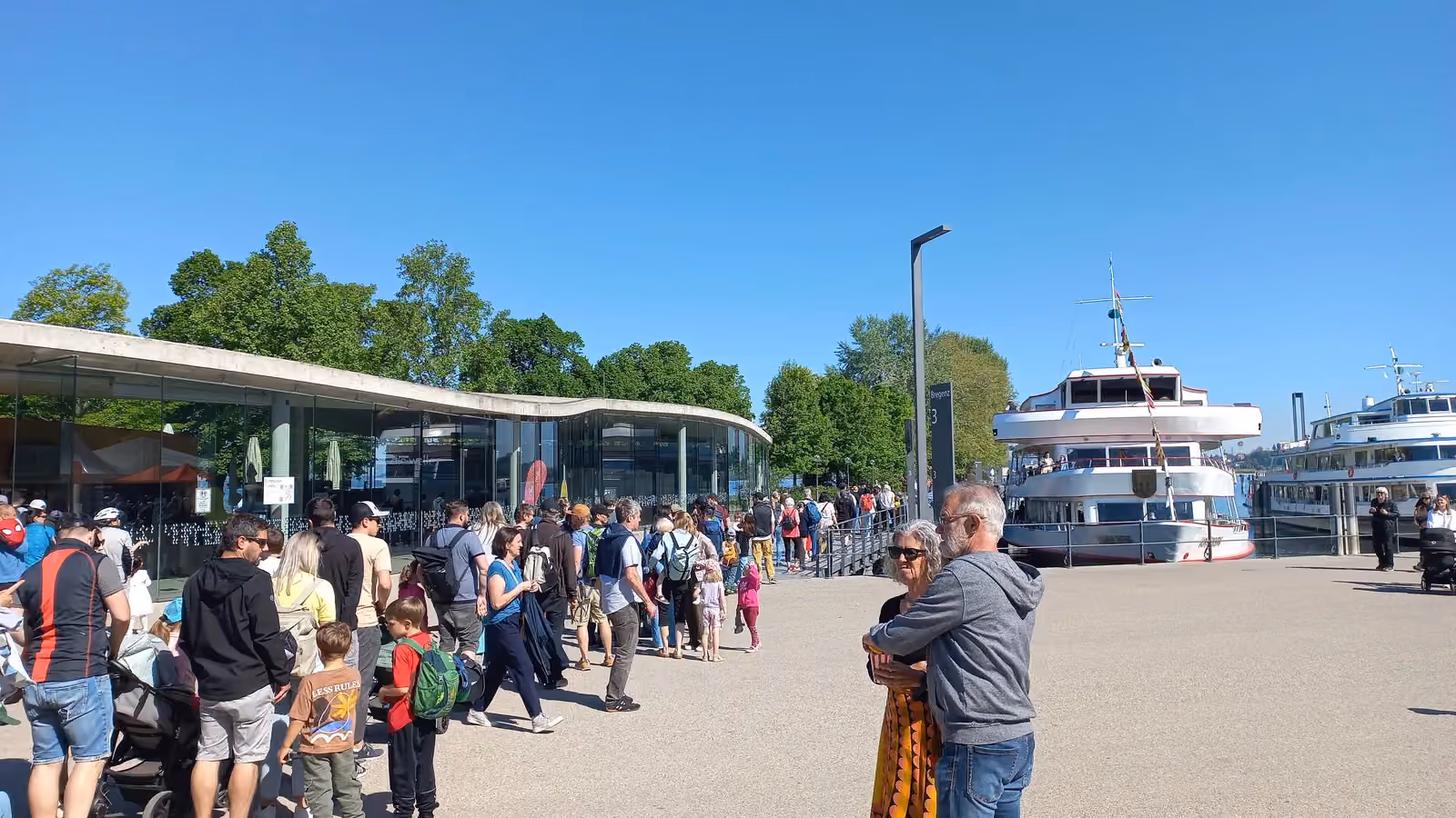 Passengers queue on a lakeside pier in Bregenz, Austria, to board a moored passenger ship; boxes and pose skeletons mark 21 people in the line.