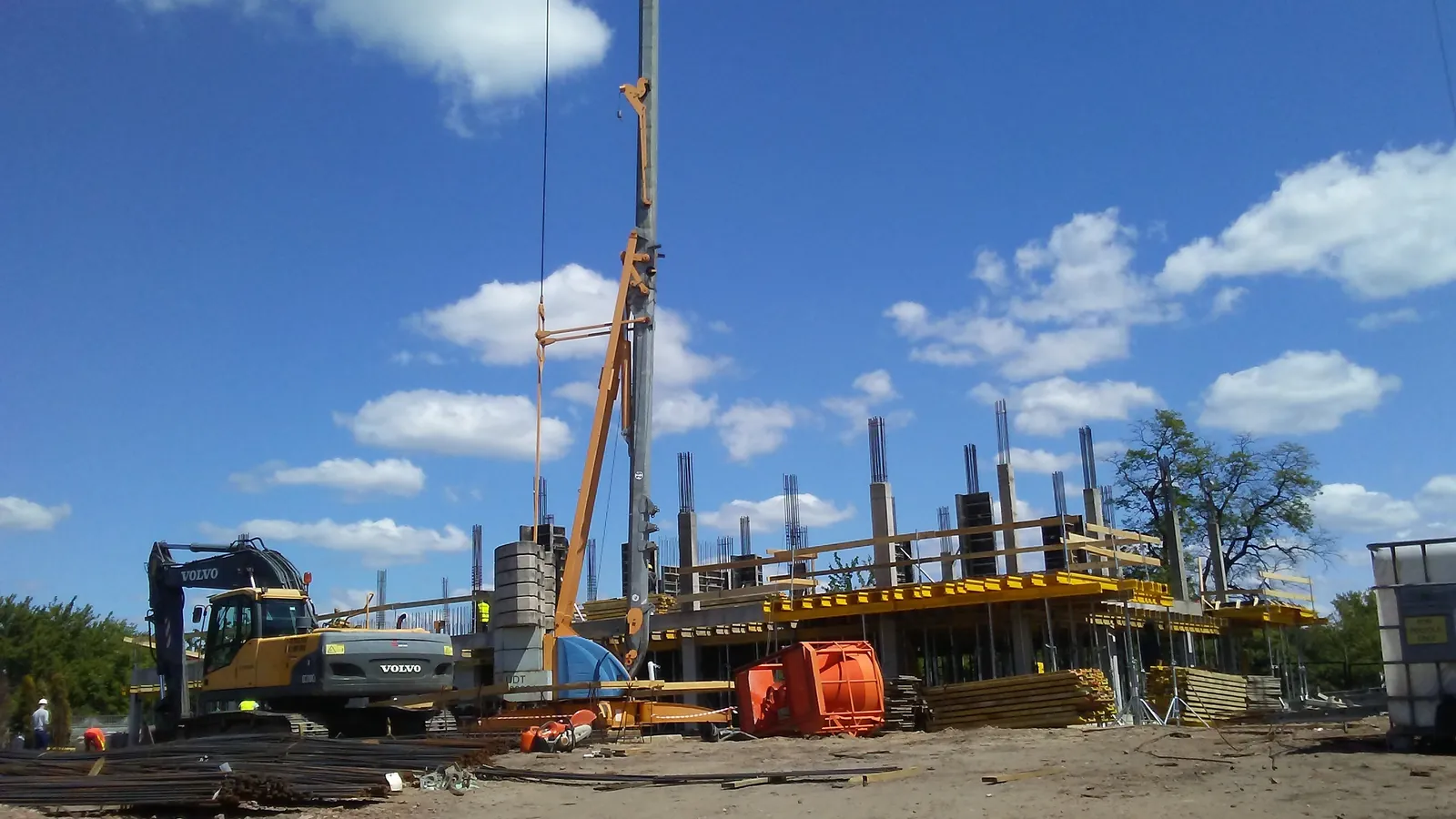 A building going up on Długa Street in Tomaszów Mazowiecki, Poland, with an excavator and a tall drilling mast beside the concrete frame; boxes mark the excavator and two workers.
