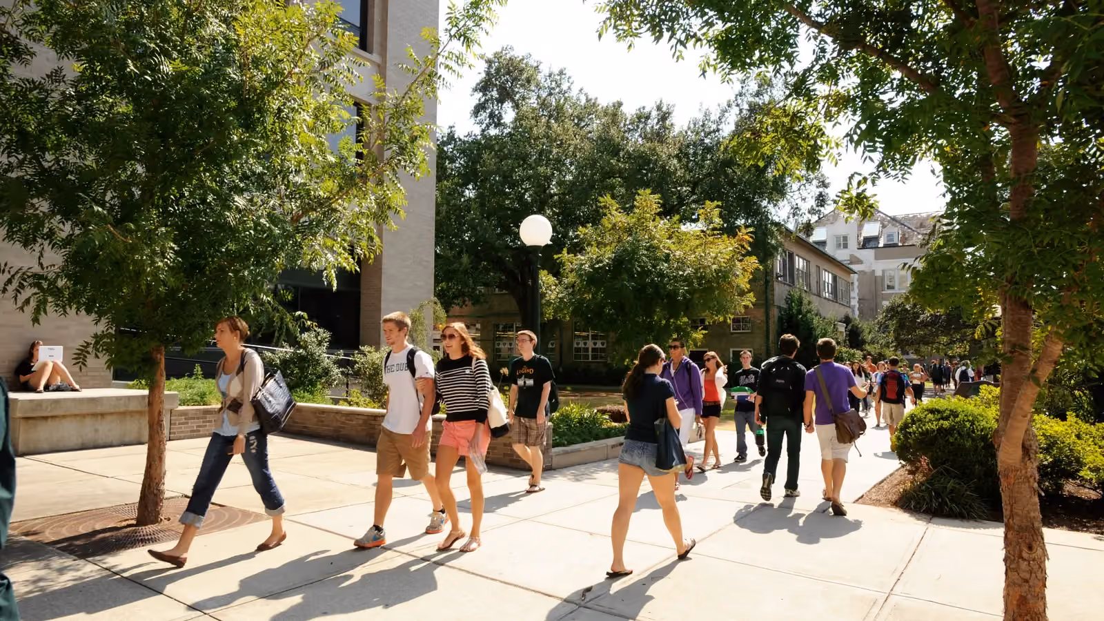 Students walk along a shaded campus walkway at Tulane University in New Orleans while one works on a laptop nearby; boxes mark 20 people and 3 backpacks.