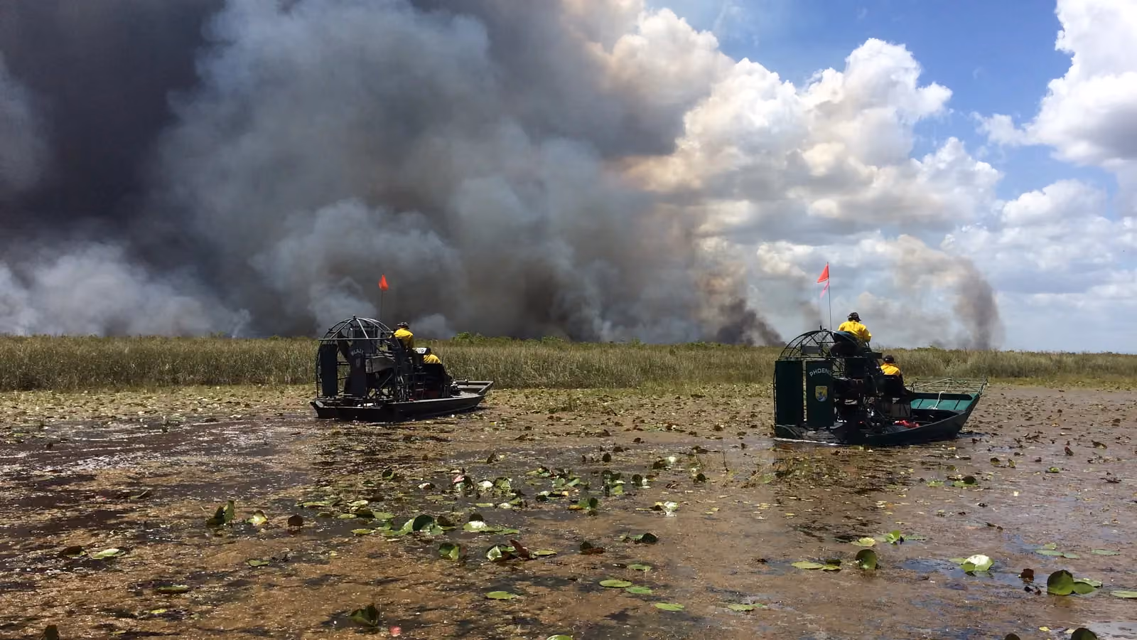 Two airboat crews patrol a lily-pad marsh at Loxahatchee National Wildlife Refuge, Florida, as smoke from a planned burn rises behind them; boxes mark two smoke plumes, both airboats and two crew members.