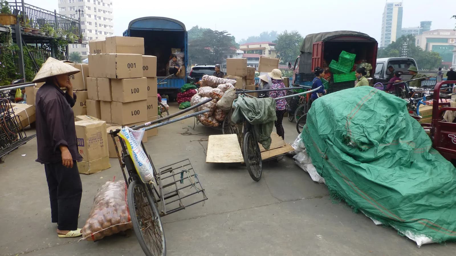 Traders with cargo bicycles unload trucks and stacked boxes outside the downtown border crossing in Hekou, China; boxes mark 11 people and 2 trucks.