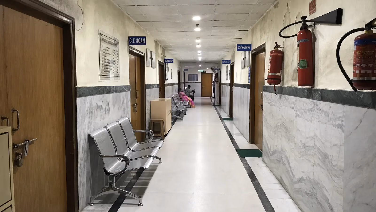 A hospital corridor lined with metal waiting benches and fire extinguishers, with a few people at the far end; boxes mark three people and two benches.