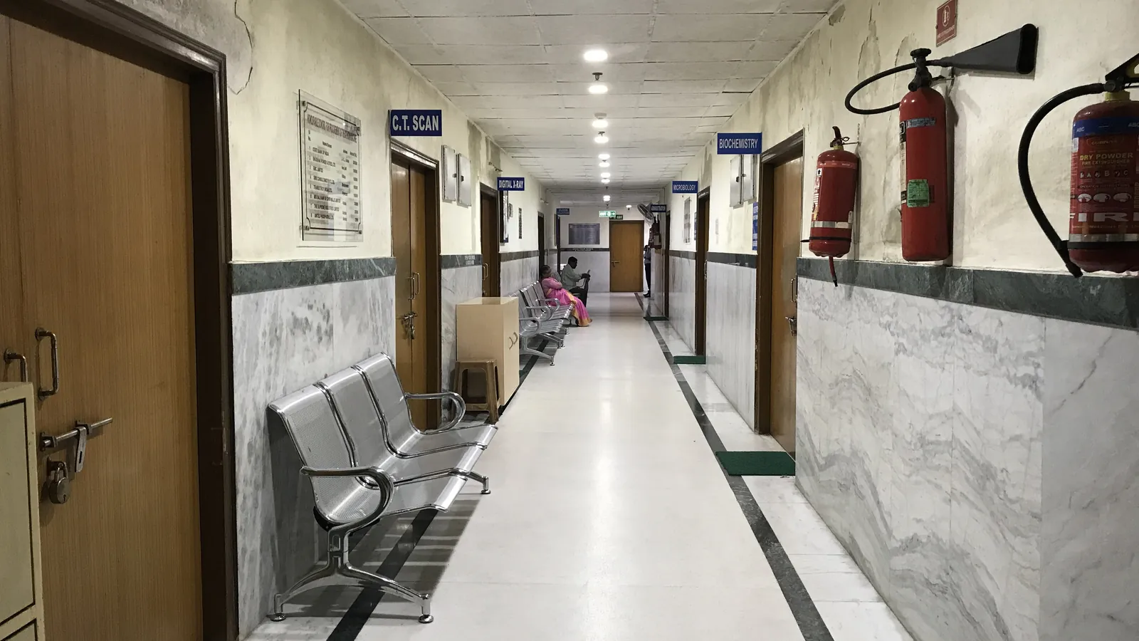 A hospital corridor lined with metal waiting benches and fire extinguishers, with a few people at the far end; boxes mark three people and two benches.