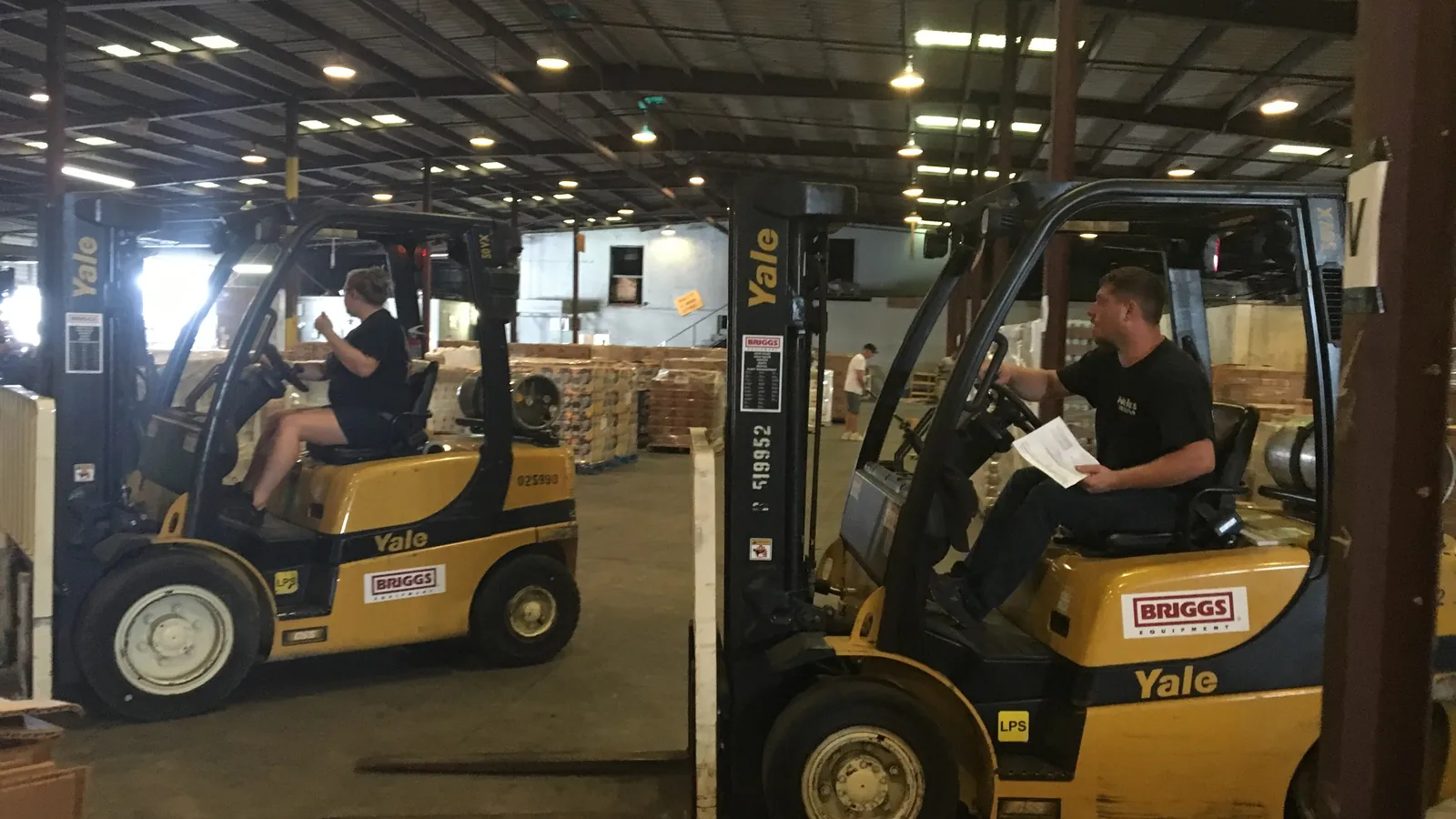 Two forklift operators driving through a warehouse in Baton Rouge, Louisiana, with a third worker in the aisle behind them; boxes mark both forklifts and all three people.
