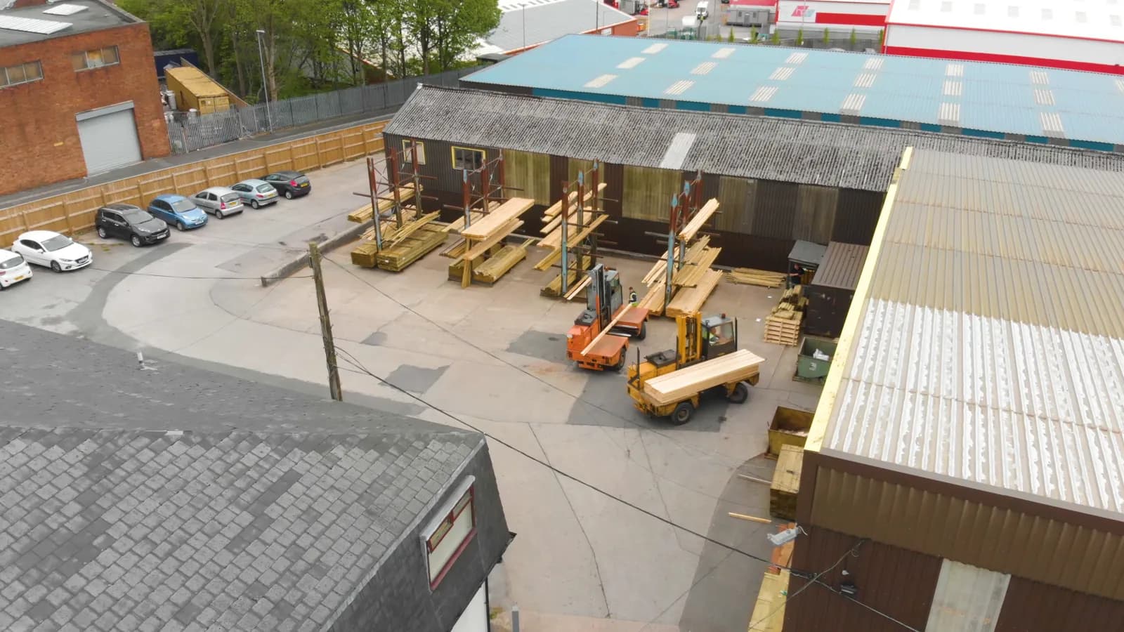 An elevated view of a timber merchant's yard where sideloader forklifts move timber between storage racks; boxes mark one forklift and two workers.