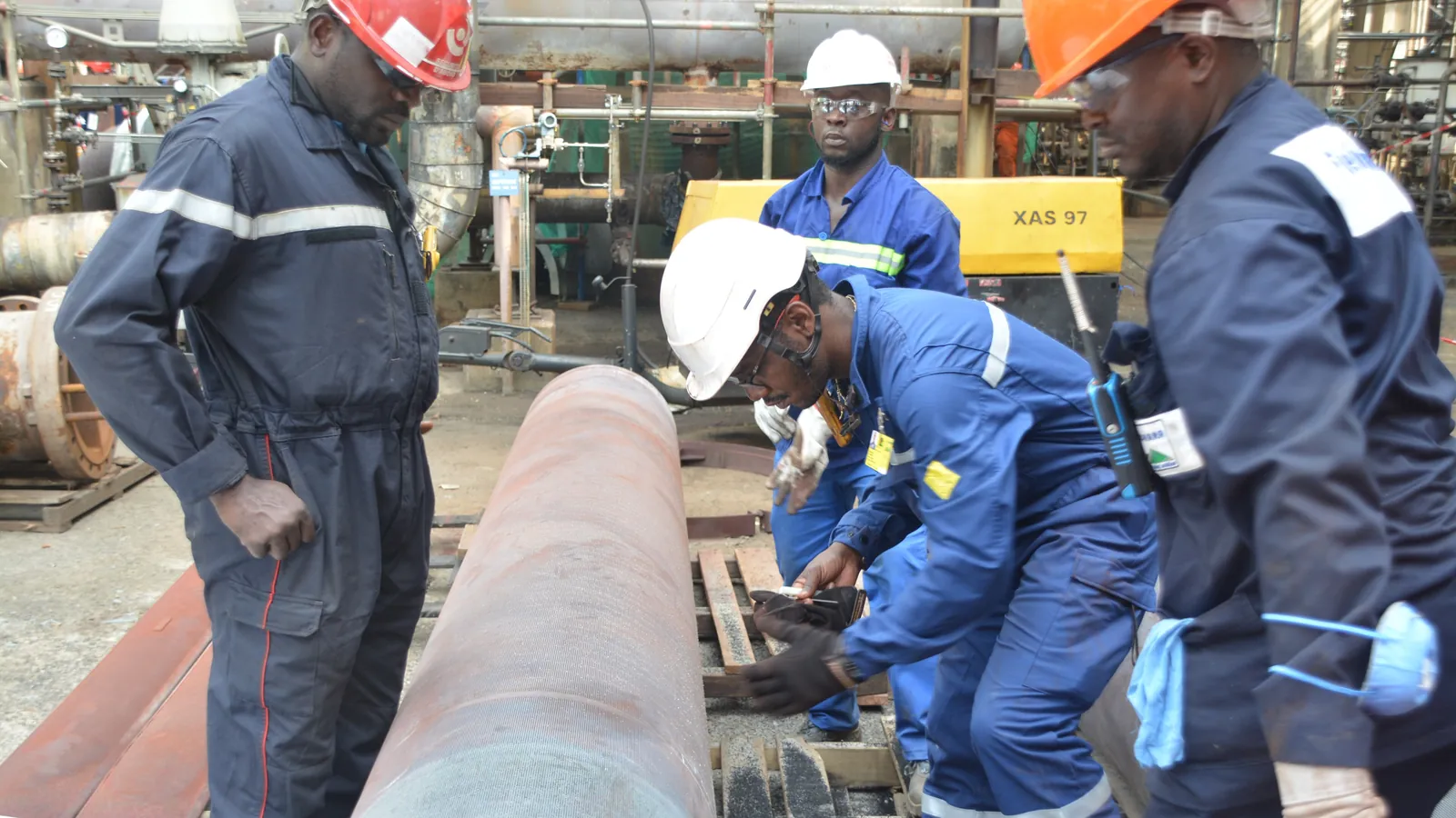 A maintenance crew in coveralls inspects a length of pipe during a refinery shutdown in Cameroon; boxes mark four workers and one hard hat.