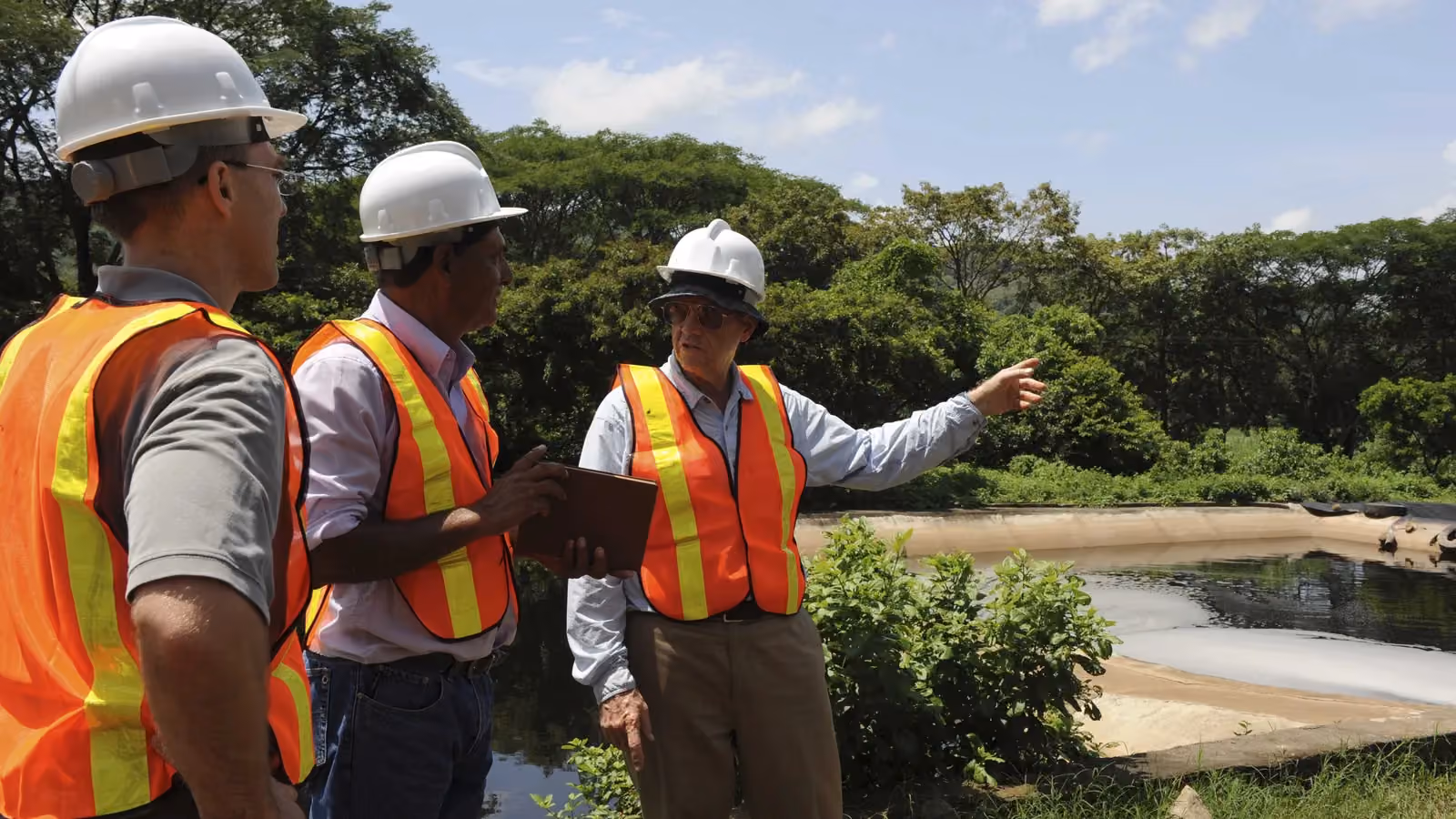 Three men in hard hats and orange safety vests inspecting a lined pond in El Salvador; boxes mark two hard hats and all three vests.