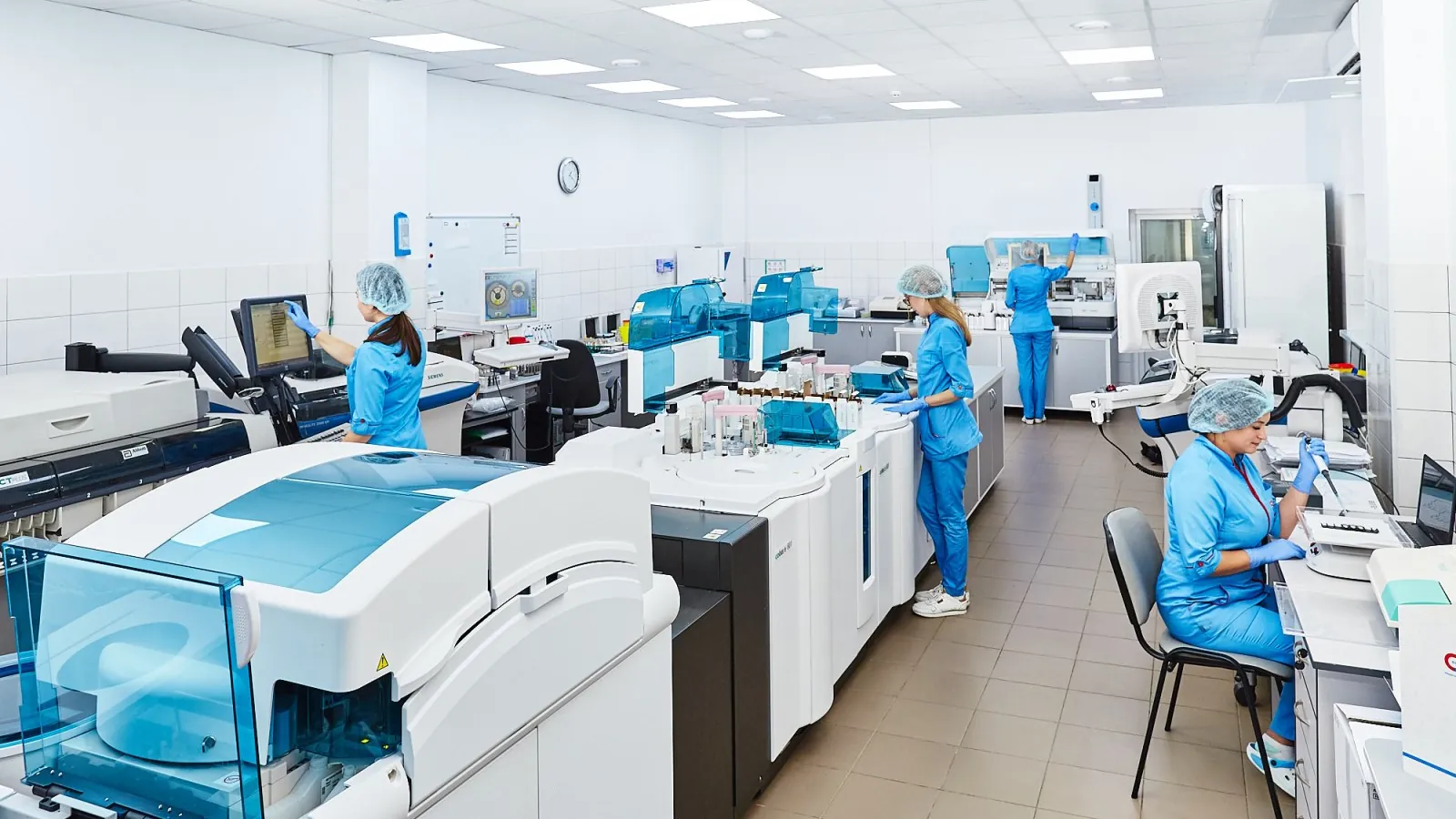 Four staff in scrubs and hair caps work at automated analyzers and computers along a lab aisle; boxes mark all four people.
