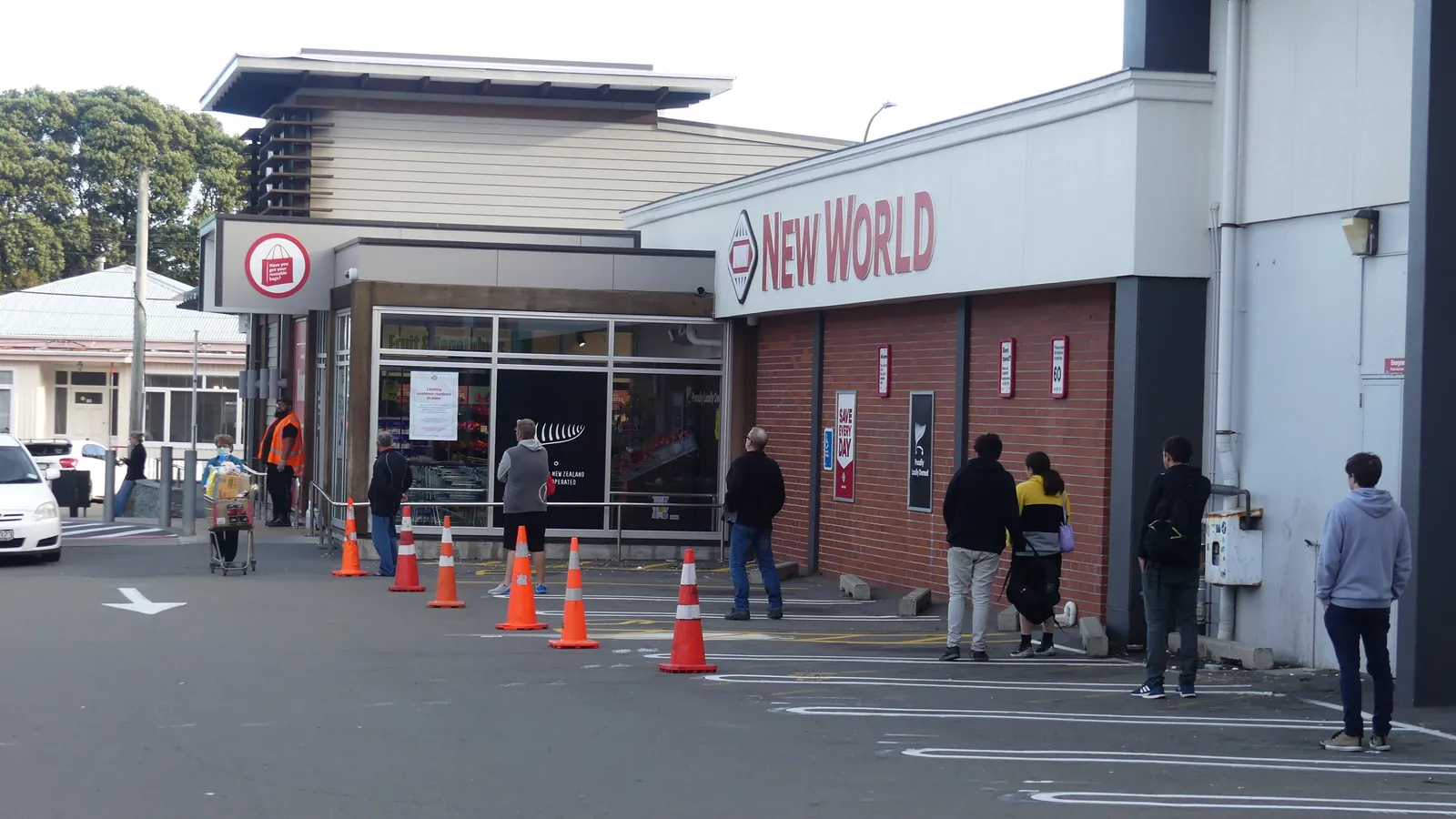 Shoppers wait in a spaced line outside a supermarket entrance; boxes mark 10 people, a bag and a shopping cart.