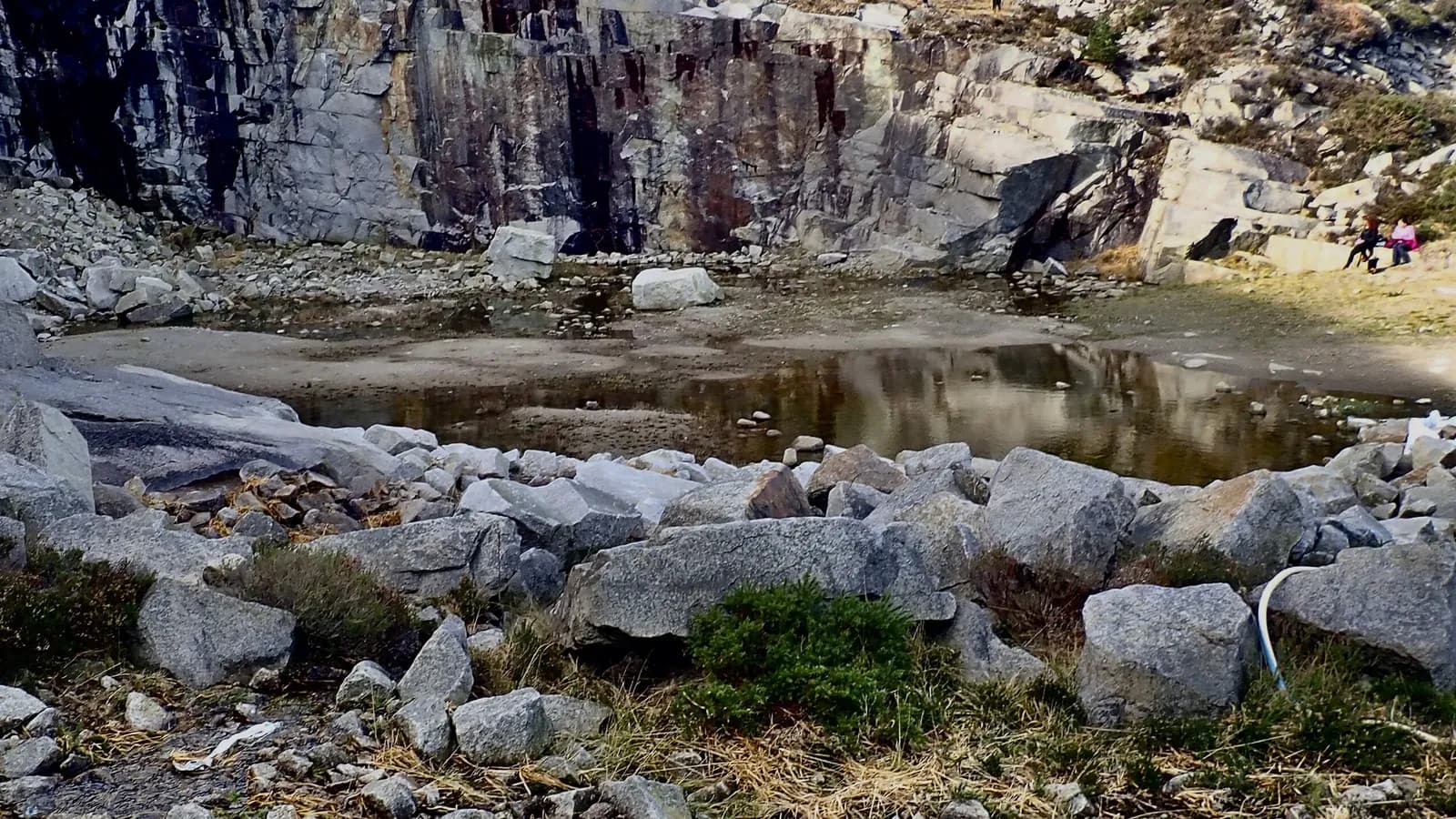 Standing water covers part of a quarry floor below a rock face; an outline traces the water, and boxes mark two people sitting on the far rocks.