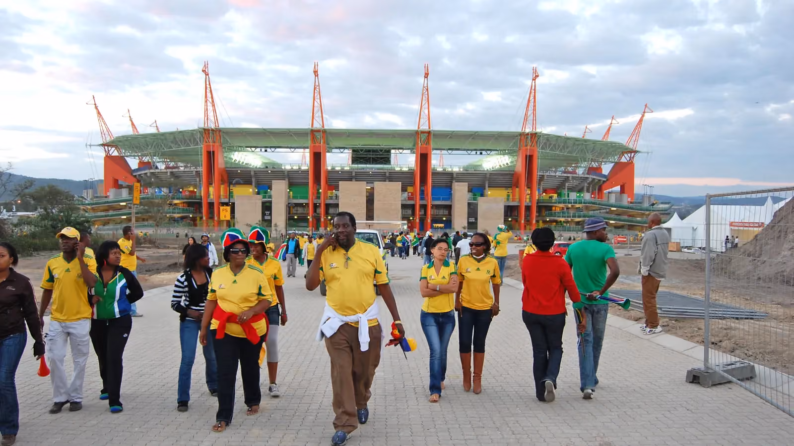 Fans in yellow team shirts walk away from Mbombela Stadium in South Africa; boxes mark 33 people.