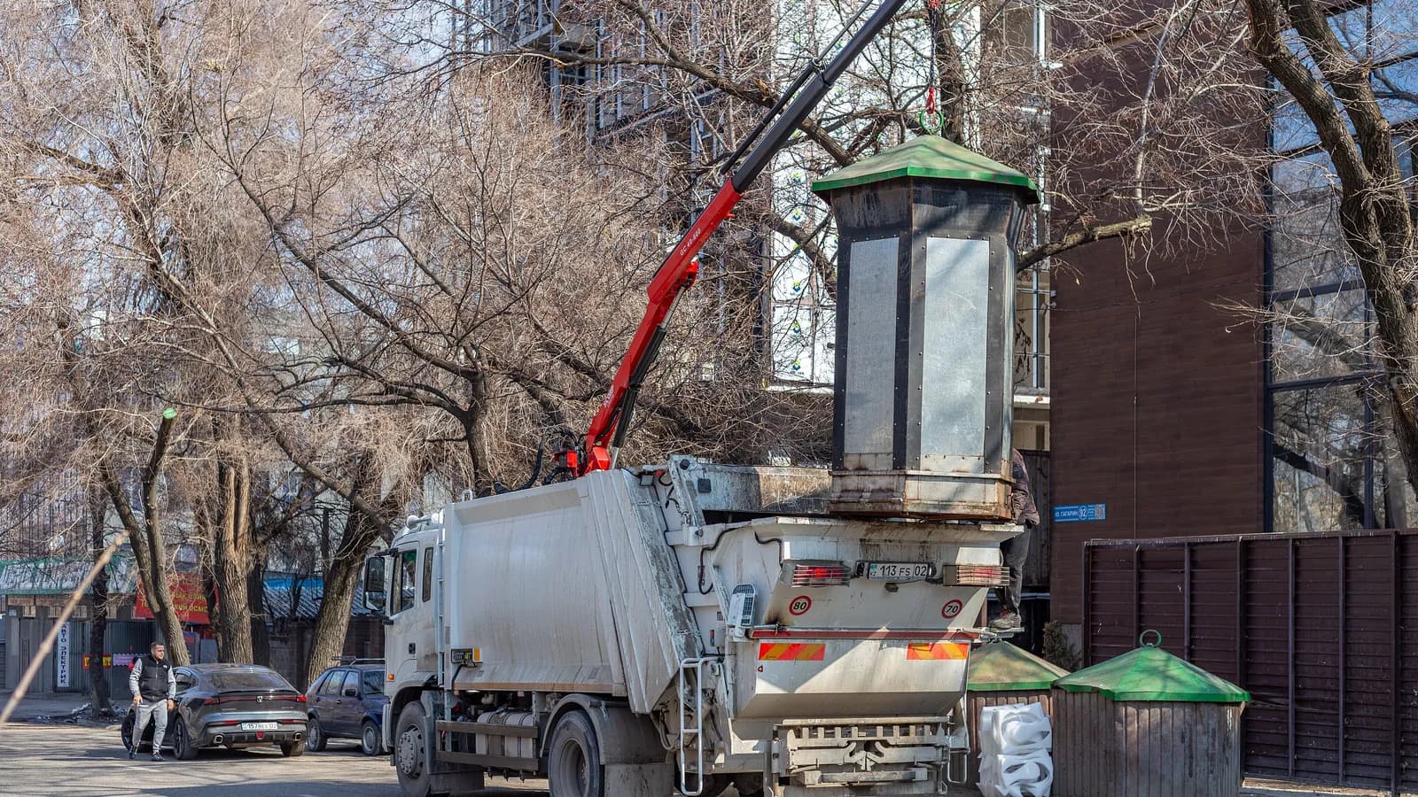 A garbage truck's crane lifts a large street bin to empty it while a man walks past; boxes mark the truck and the passerby.