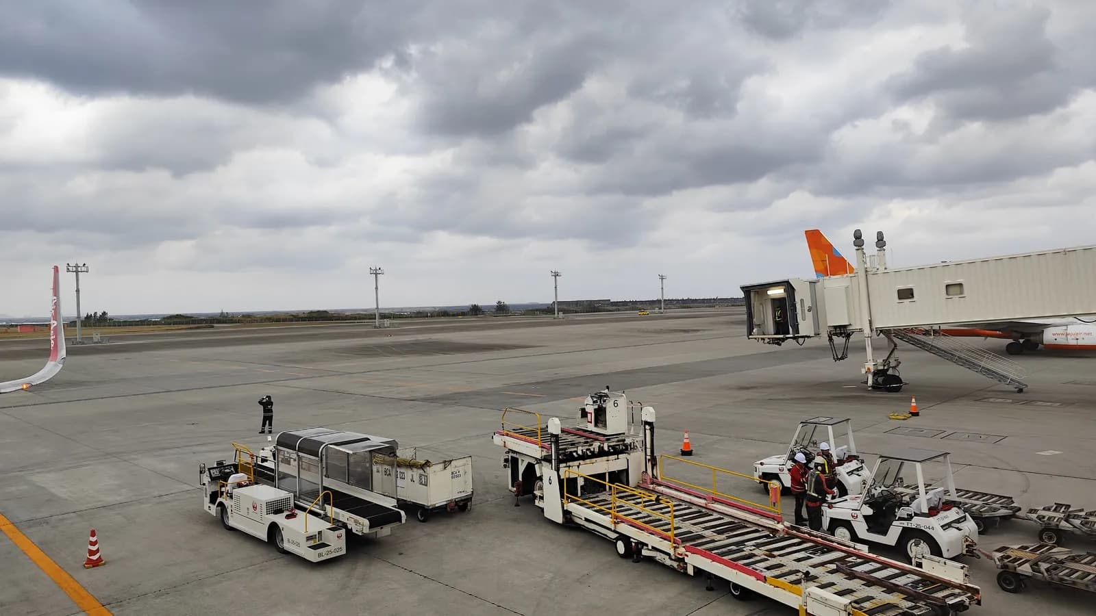 Ground crew work beside a cargo loader and baggage tugs near a jet bridge on the apron at Naha Airport in Okinawa; boxes mark 2 aircraft, 4 ground service vehicles and 4 workers.