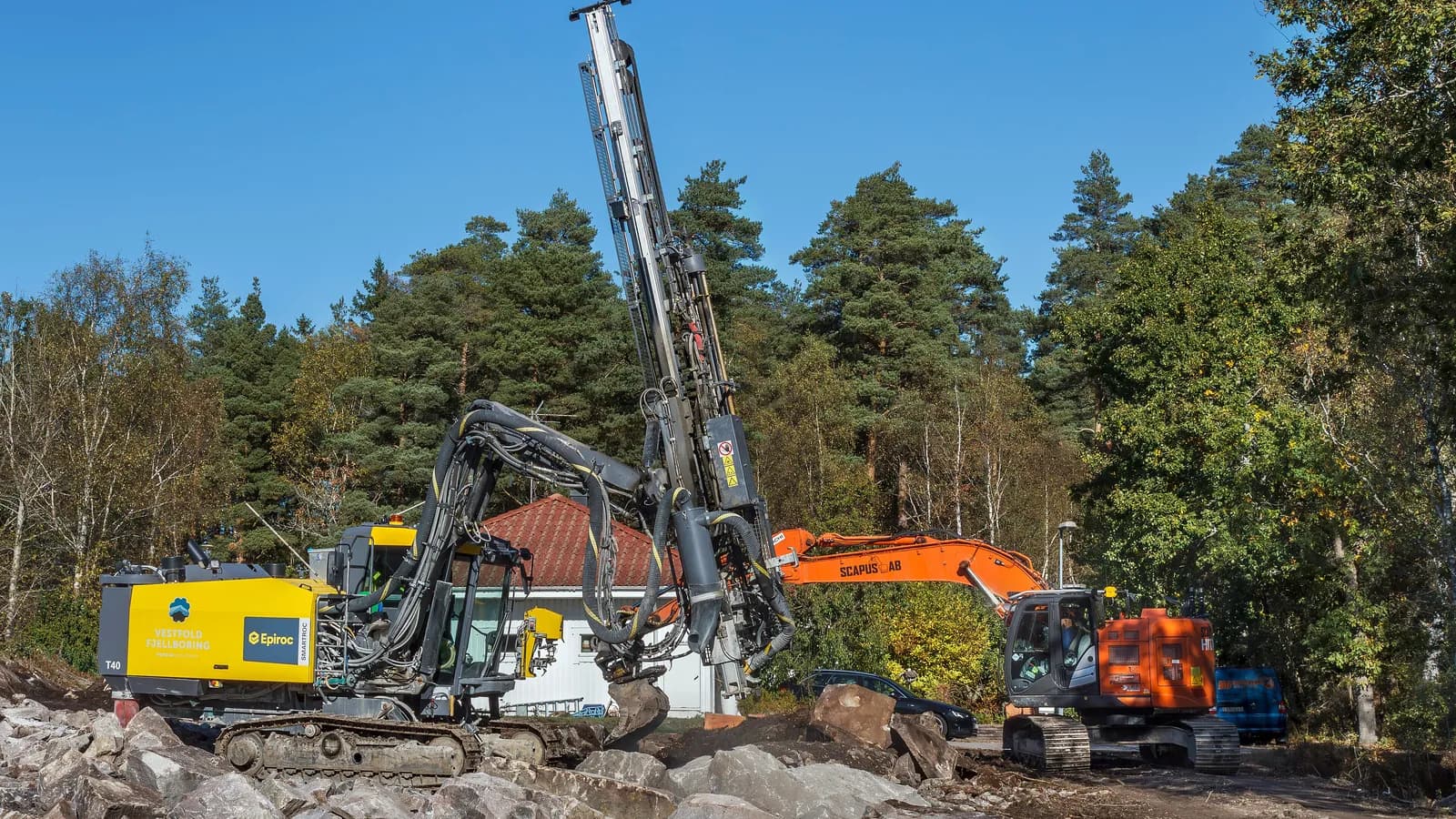 A house construction site in Tuntorp where a surface drill rig bores into granite; boxes mark an excavator and a parked car.