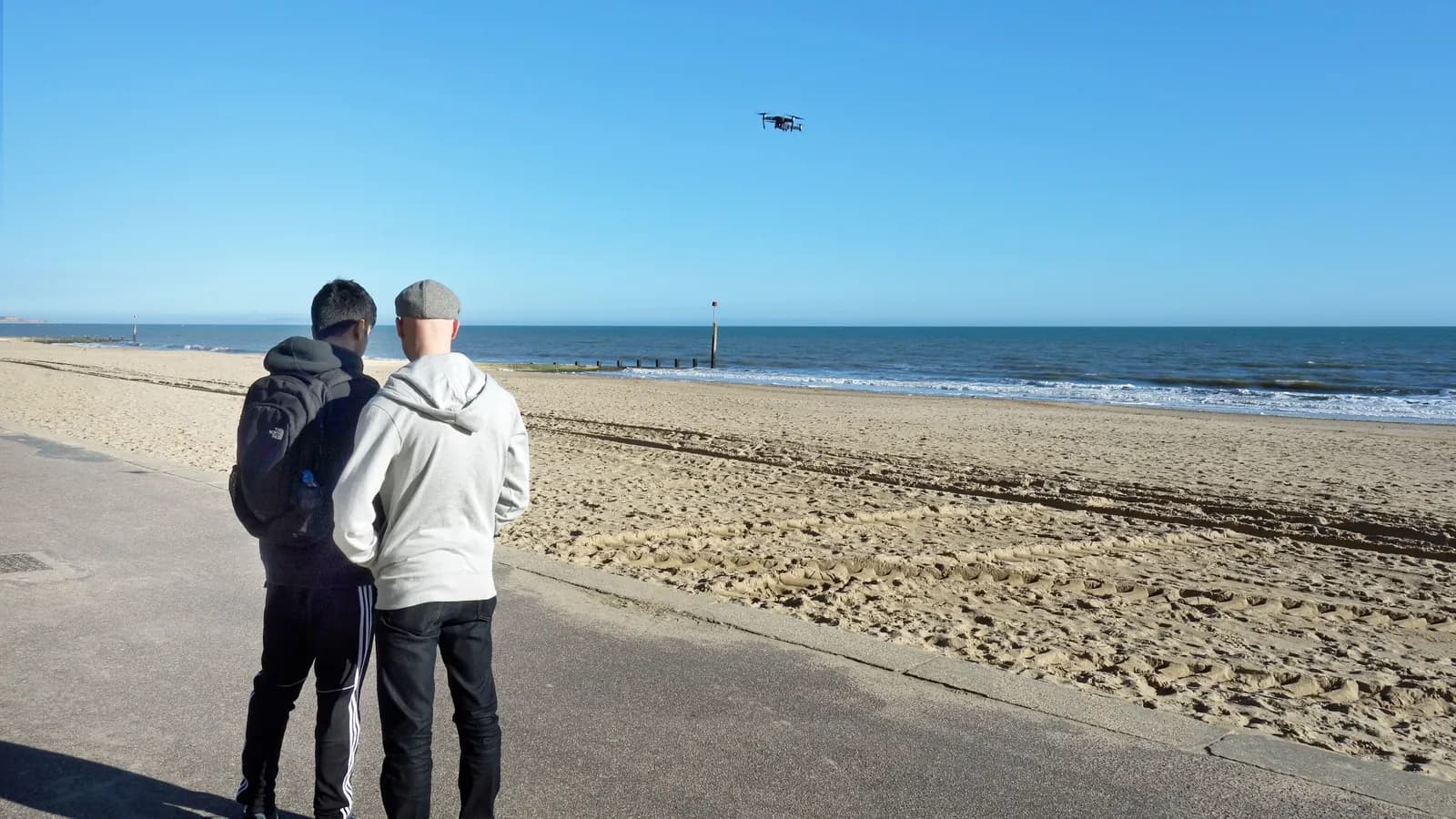 Two people on a beach promenade fly a small quadcopter over the sand; boxes mark the drone in the sky and both people.