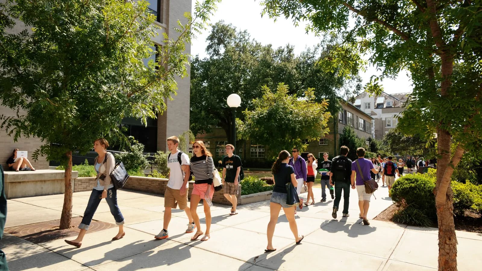 Students walk along a shaded campus walkway at Tulane University in New Orleans while one works on a laptop nearby; boxes mark 20 people and 3 backpacks.