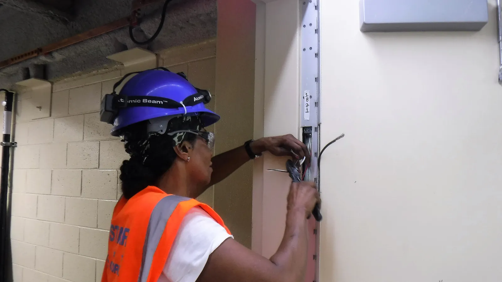 A worker in a blue hard hat and orange safety vest wiring an access control panel inside a power substation in Queens, New York; boxes mark the worker, her hard hat and her vest.