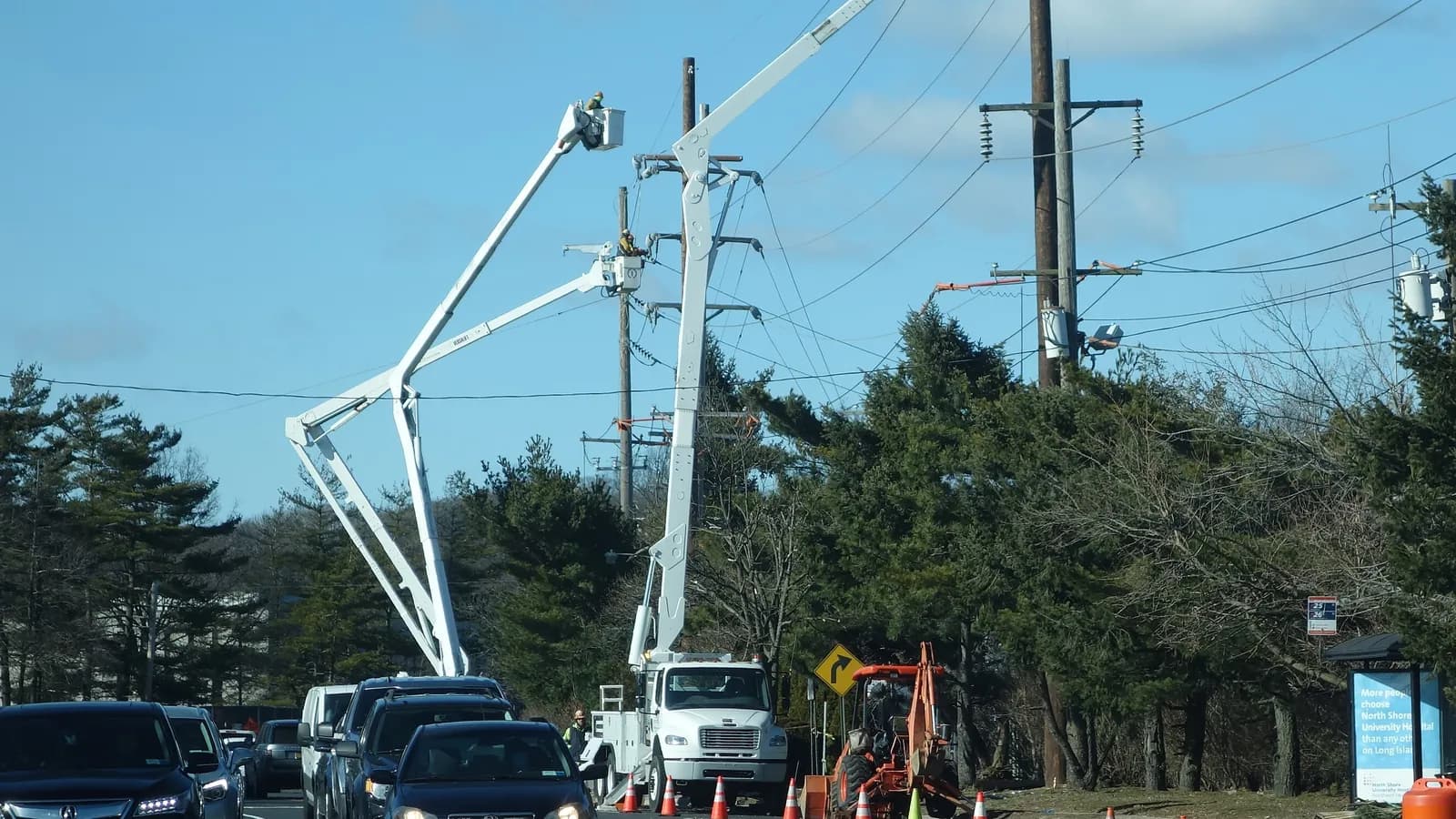Line workers in bucket trucks work on power lines above Lakeville Road at the edge of Queens, New York; boxes mark a bucket truck, a worker beside it and the hard hat of a worker in a raised bucket.