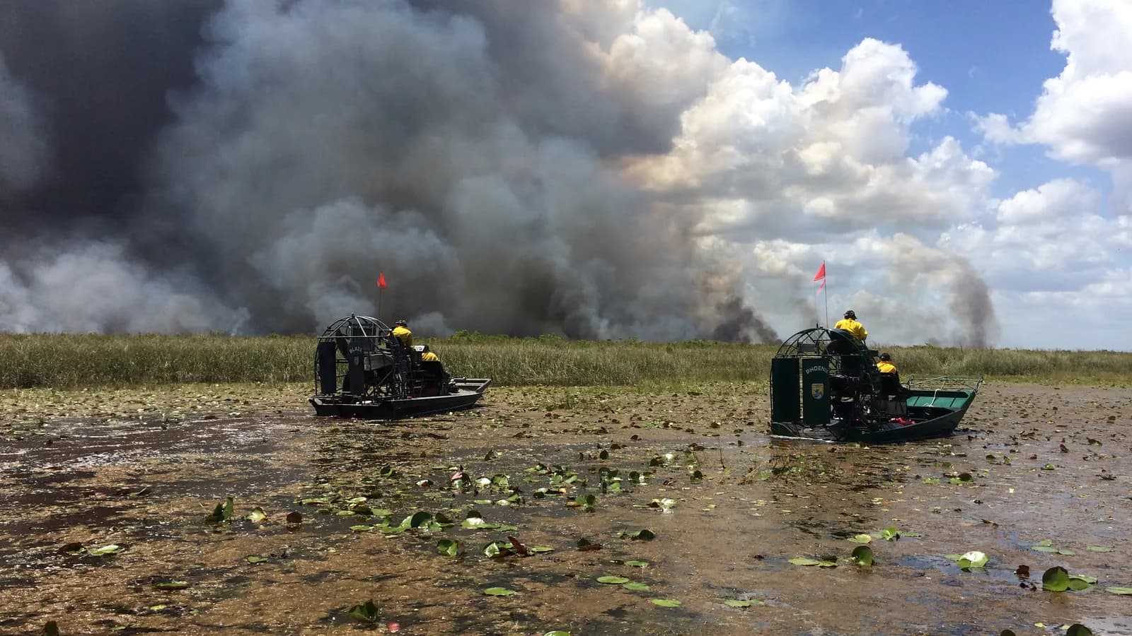 Two airboat crews patrol a lily-pad marsh at Loxahatchee National Wildlife Refuge, Florida, as smoke from a planned burn rises behind them; boxes mark two smoke plumes, both airboats and two crew members.