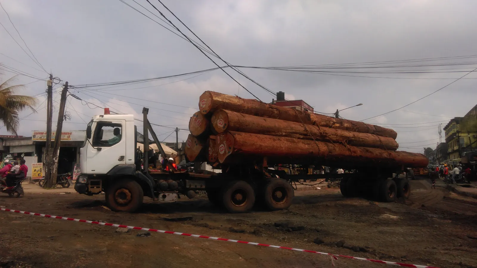 A logging truck loaded with large tropical logs passing through a town street; boxes mark the truck, its driver and nine people nearby.