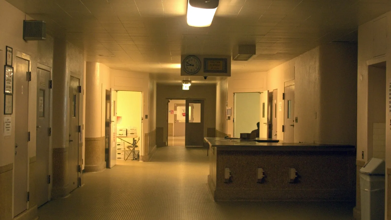 A dimly lit hallway in an old hospital building, with one person seated behind a long counter; boxes mark the person and a chair.