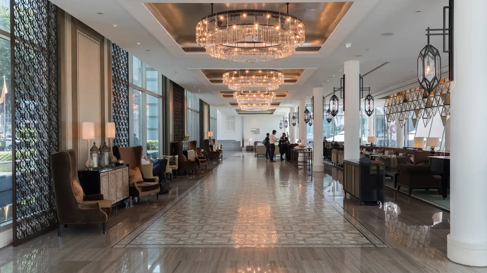 Armchairs line a long hotel lobby lounge under crystal chandeliers as guests gather at the far end; boxes mark 8 chairs and 6 people.