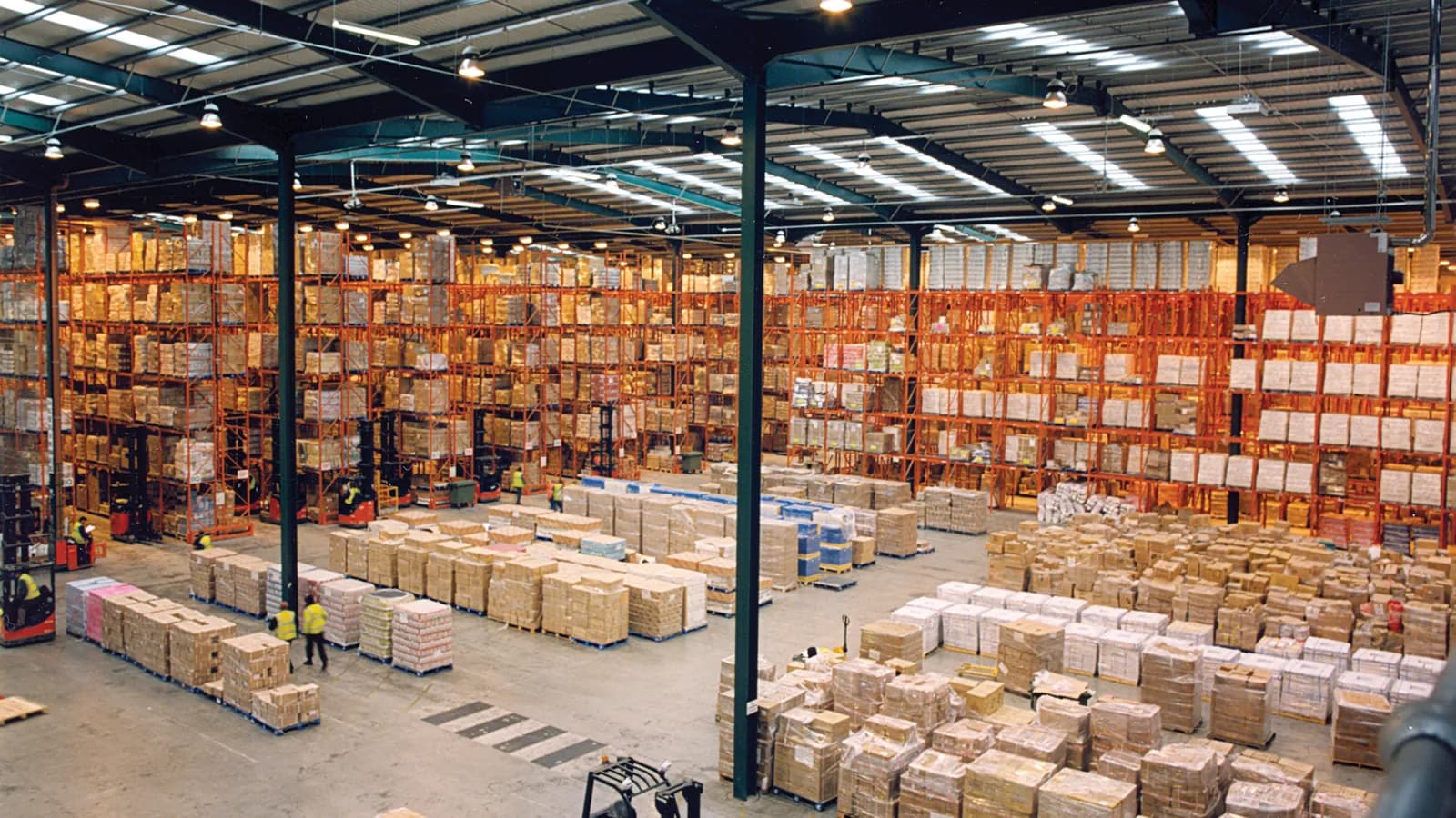 An elevated view across a large warehouse with tall orange pallet racking and rows of stacked goods; boxes mark six workers on the floor.