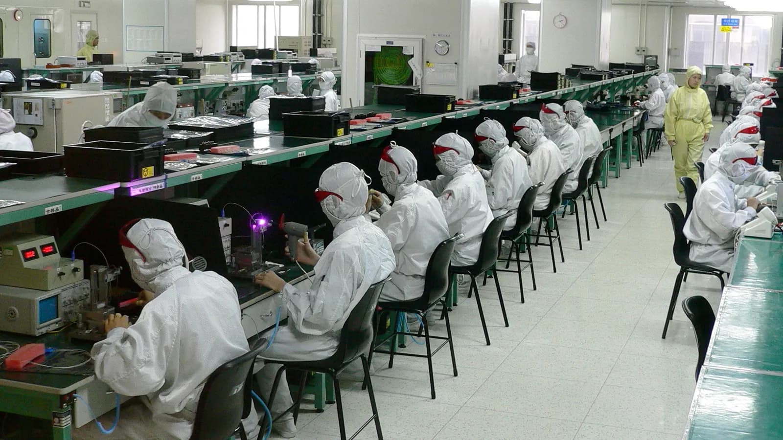 Workers in white cleanroom suits assemble components at long benches in an electronics factory in Shenzhen, China; boxes mark 20 workers.