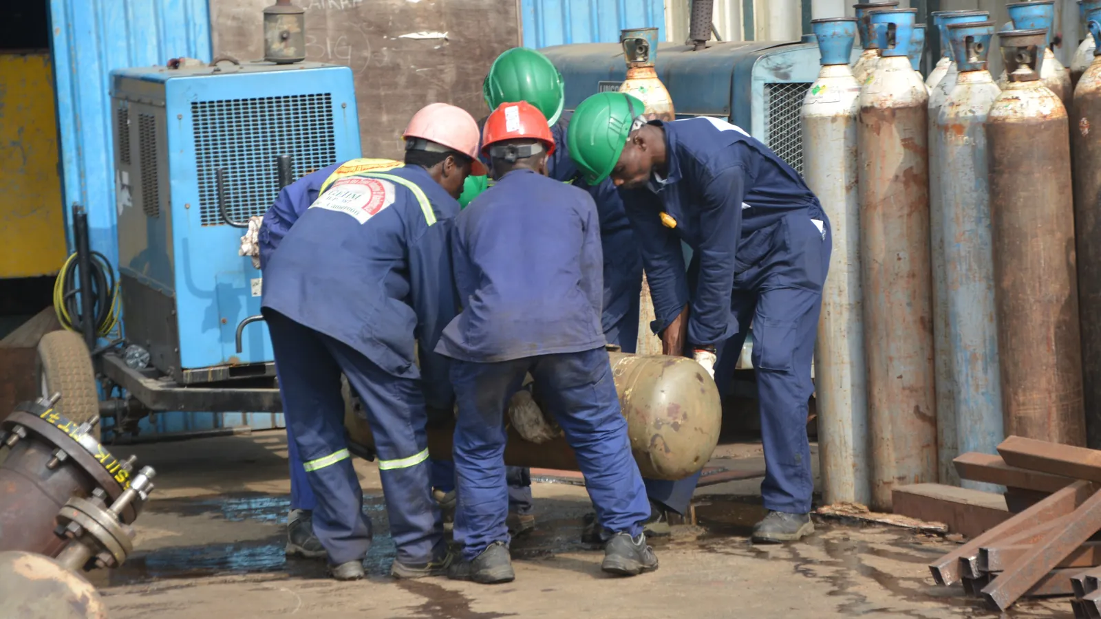 Workers in hard hats lift a metal vessel beside rows of gas cylinders at a refinery in Cameroon; boxes mark three workers and two hard hats.