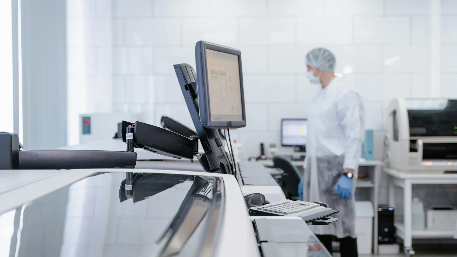 A technician in a gown, hair cover, face mask and gloves stands among lab instruments; boxes mark the technician and the face mask.