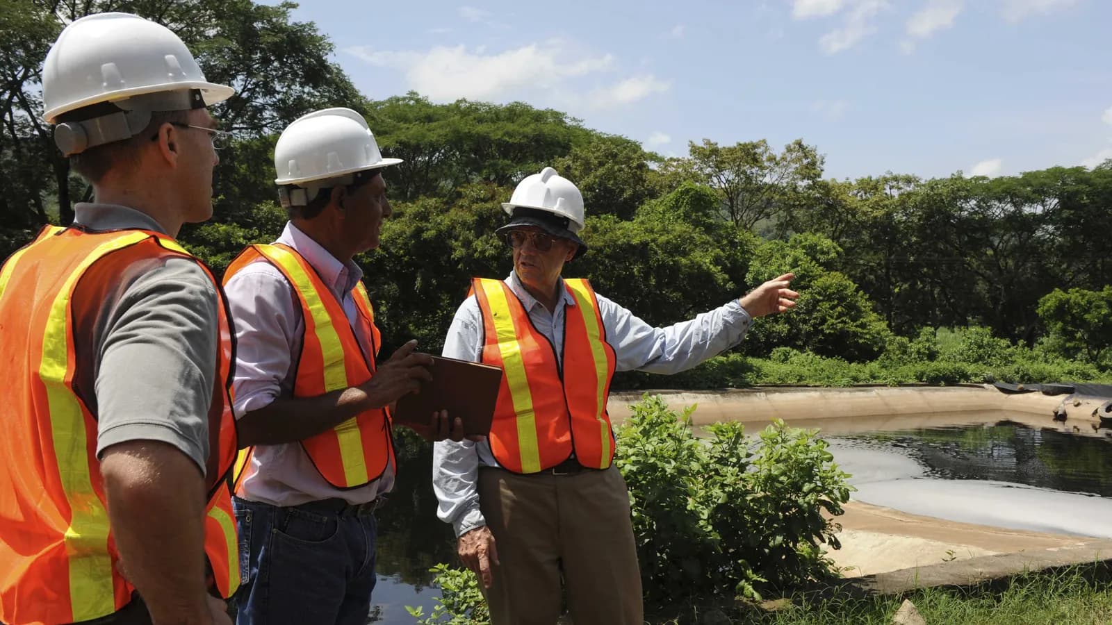 Three men in hard hats and orange safety vests inspecting a lined pond in El Salvador; boxes mark two hard hats and all three vests.