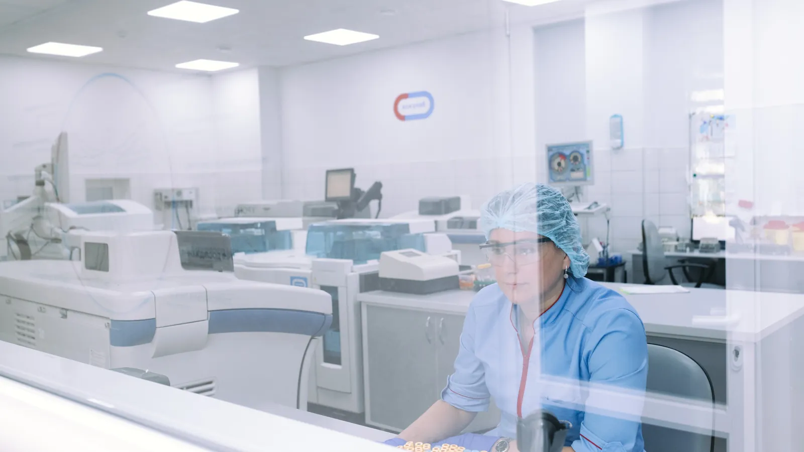 Seen through a glass partition, a technician in a hair cap and safety glasses sits at a bench among lab analyzers; a box marks her.