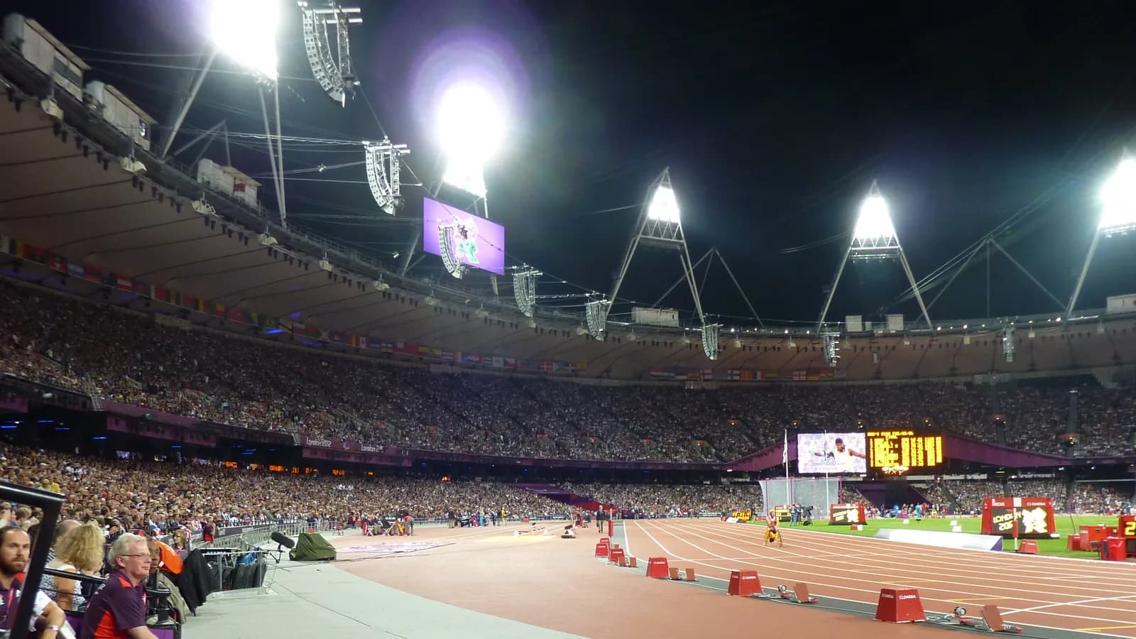 Spectators fill the stands under floodlights during an evening athletics meet; boxes mark 15 people in the nearest rows and beside the track.