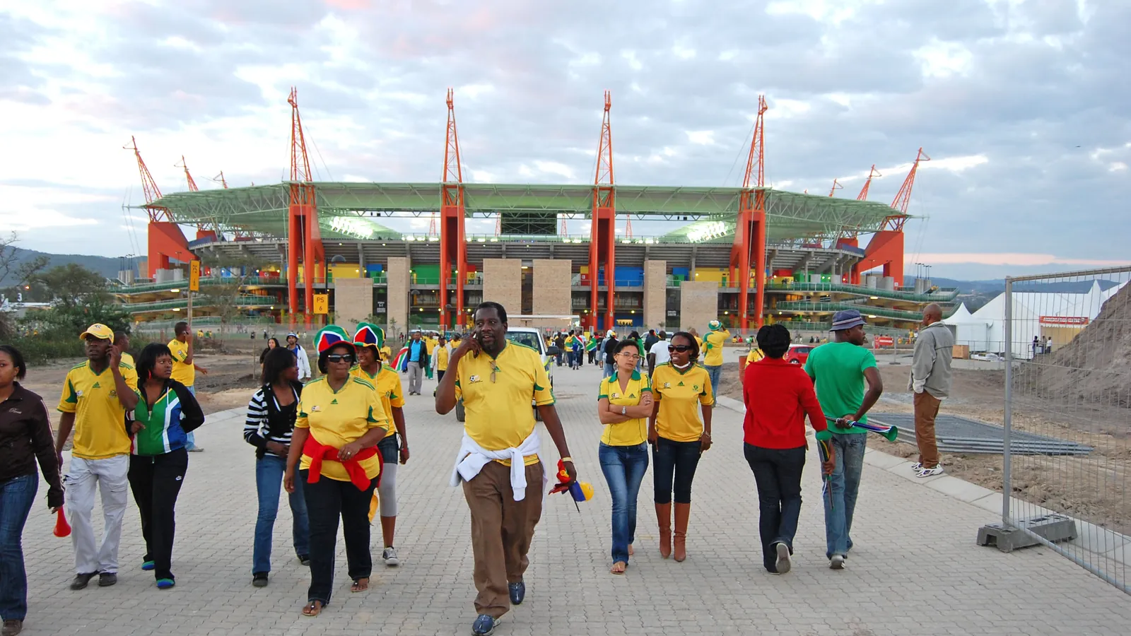 Fans in yellow team shirts walk away from Mbombela Stadium in South Africa; boxes mark 33 people.