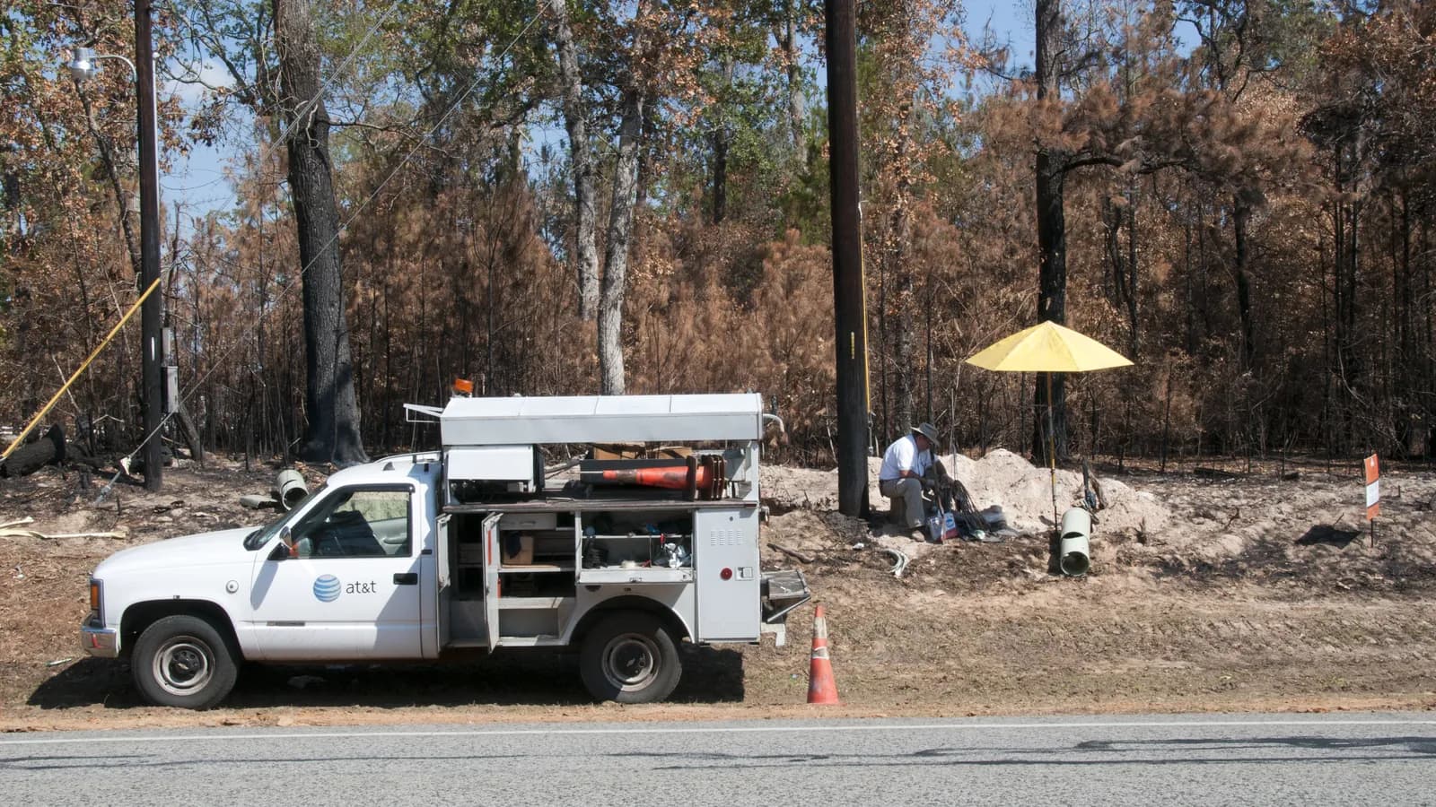 A telephone repairman works on cables at the roadside beside his service truck while restoring phone service after the 2011 wildfires near Magnolia, Texas; boxes mark the truck and the repairman.