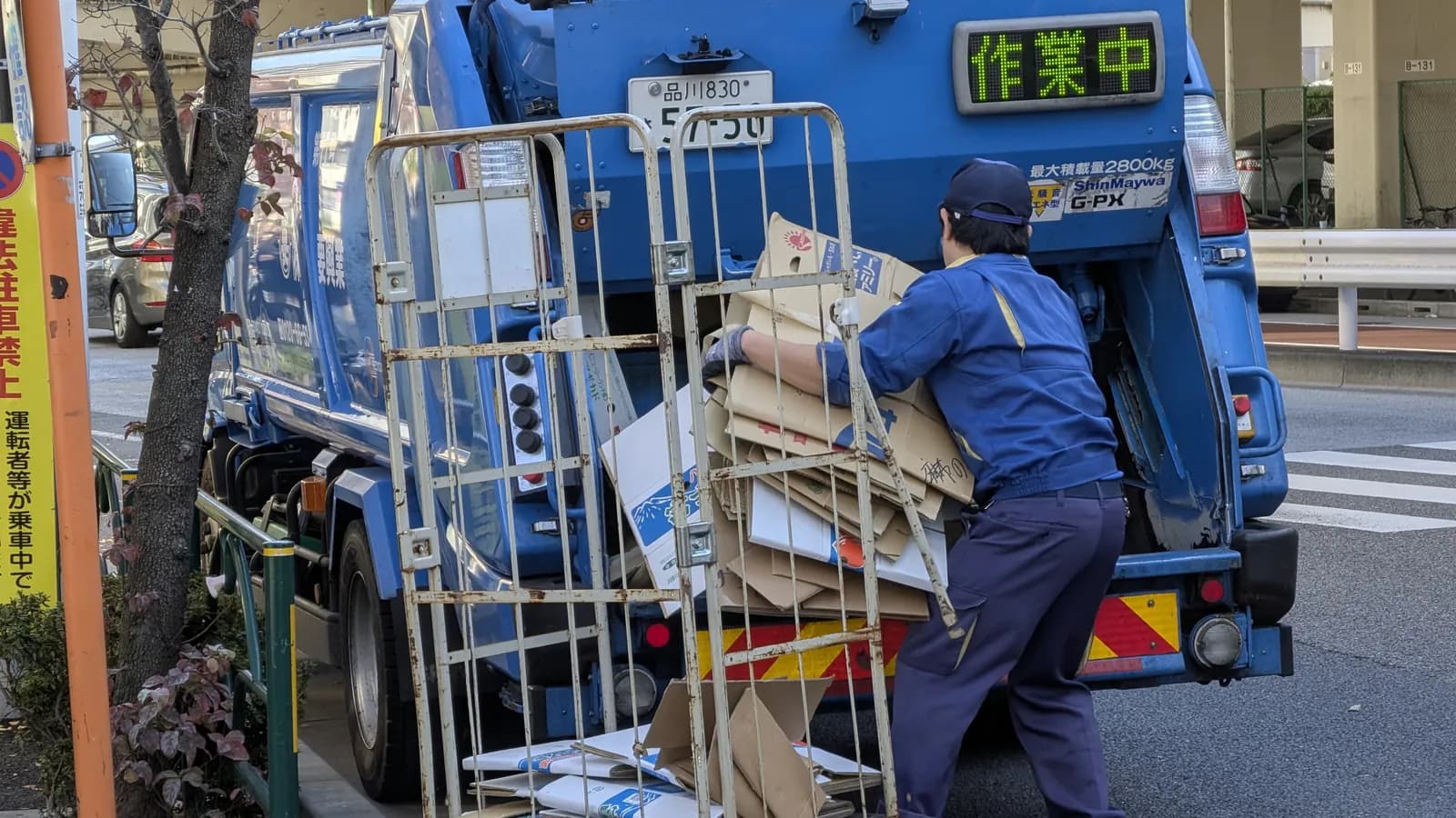 A worker pushes flattened cardboard from a wire cart into the back of a garbage truck; boxes mark the worker and the truck.