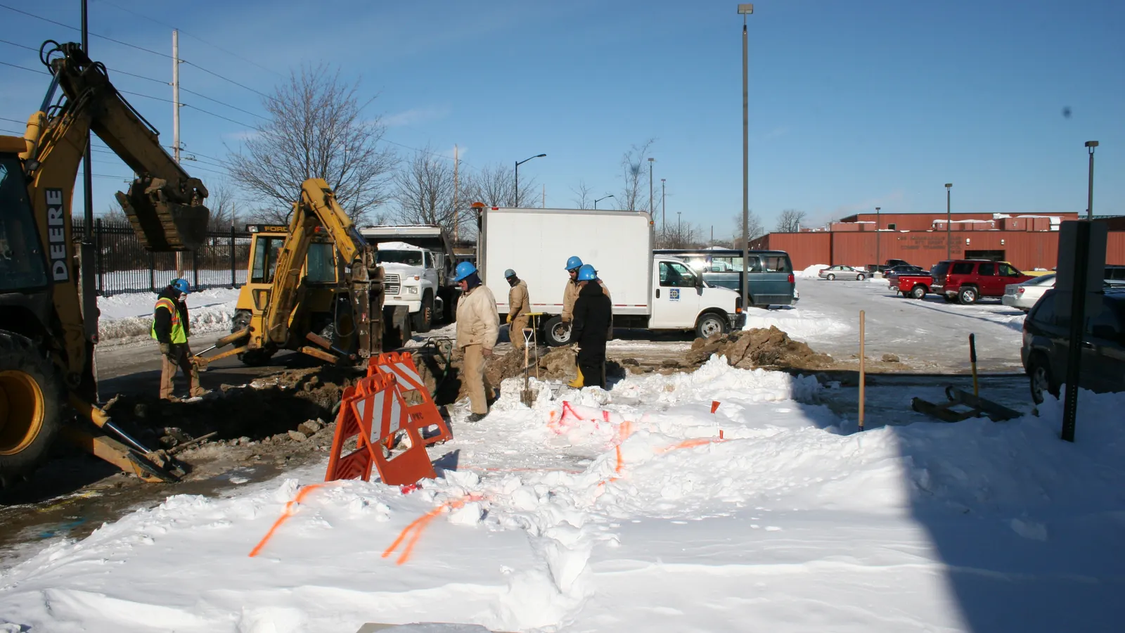 A crew digs out a broken water main in the snow beside backhoes and work trucks; boxes mark five workers, two trucks, two hard hats and a safety vest.